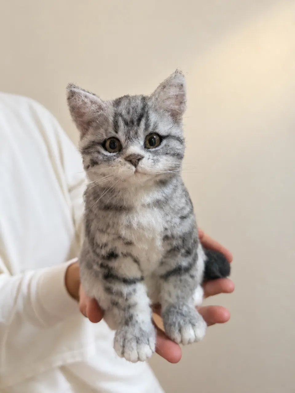 A needle-felted grey tabby kitten sitting upright, held in two hands, with striped markings and pale green eyes.