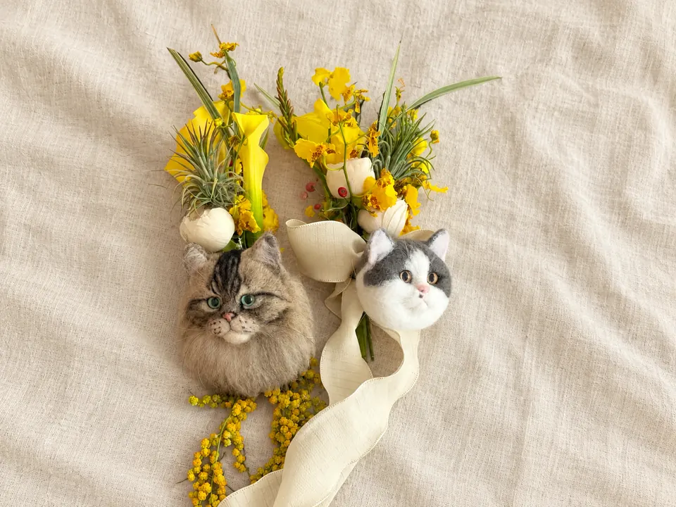 Two needle-felted cat head accessories laid on linen with yellow mimosa sprigs and cream ribbons.