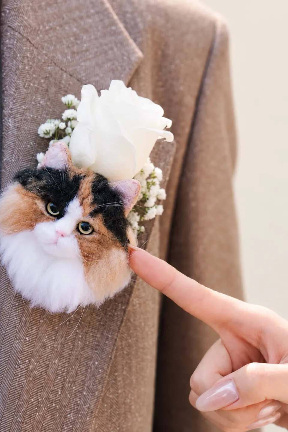 A close-up of a needle-felted calico cat corsage and white flowers pinned to a taupe jacket, with a fingertip beside it and no face visible.