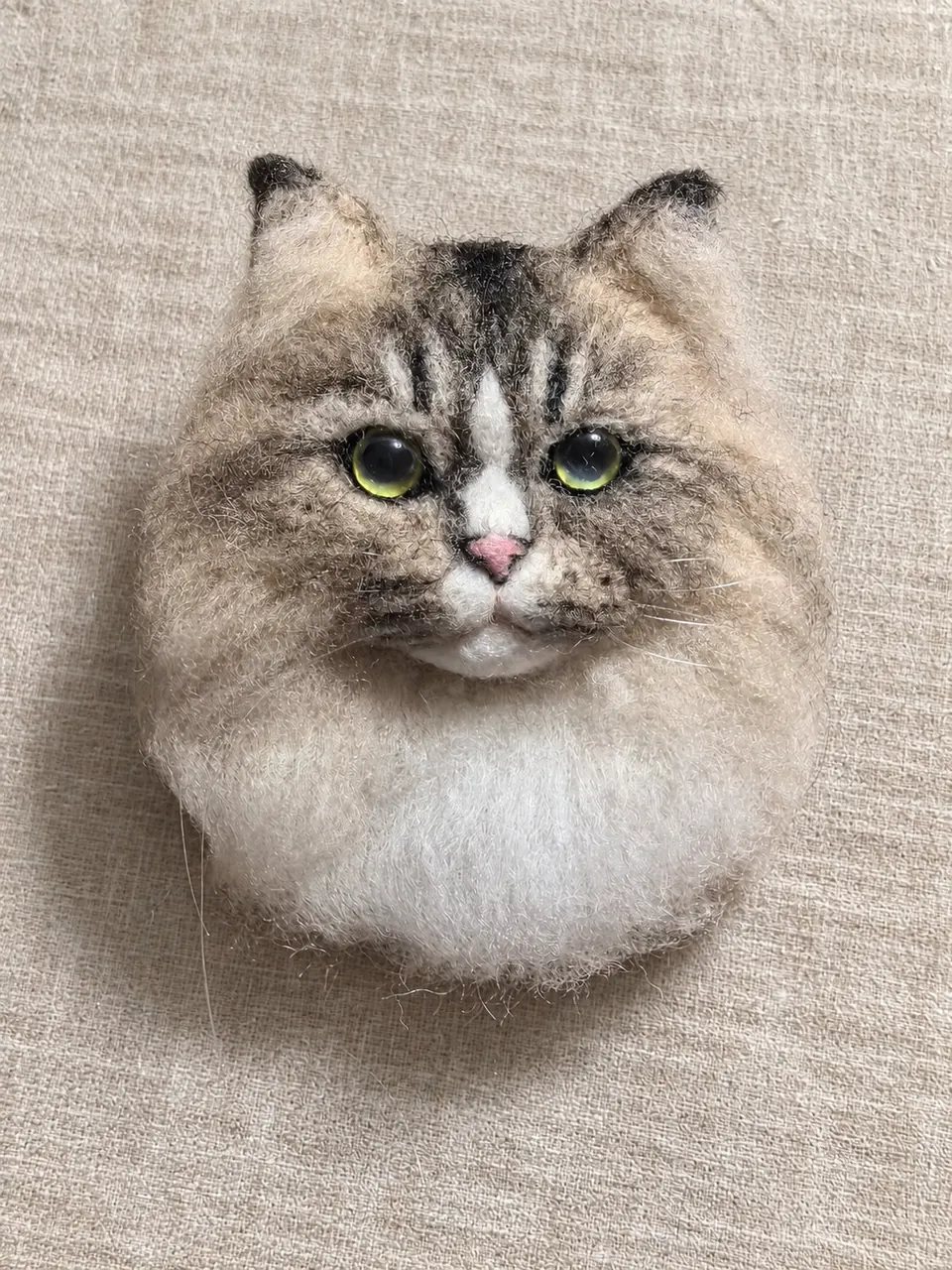 A needle-felted grey-and-white cat face portrait on oatmeal linen, with long fur worked out around the cheeks.