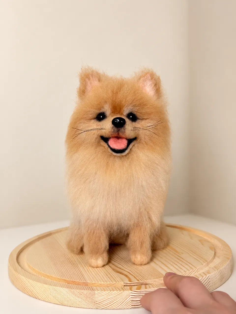 An orange needle-felted Pomeranian standing on a round wooden tray, with a fluffy coat and pink tongue, against a warm ivory background.